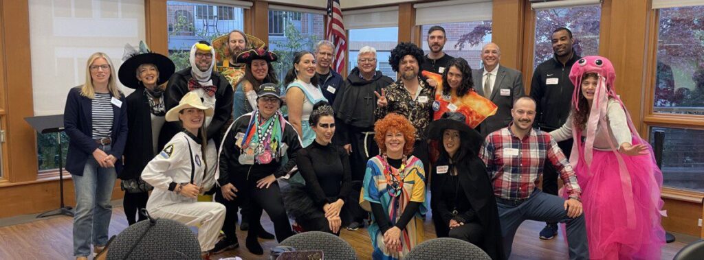 A diverse group of people in various costumes, including a witch, astronaut, penguin, and jellyfish, pose for a photo indoors.