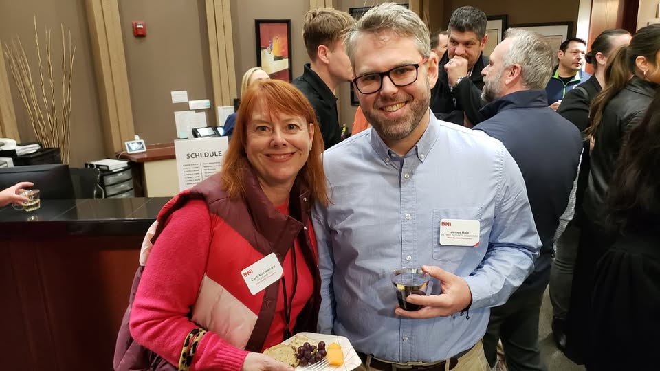 Two BNI members smile at a networking event, one holding a plate of food and the other a drink, with other attendees visible in the background.