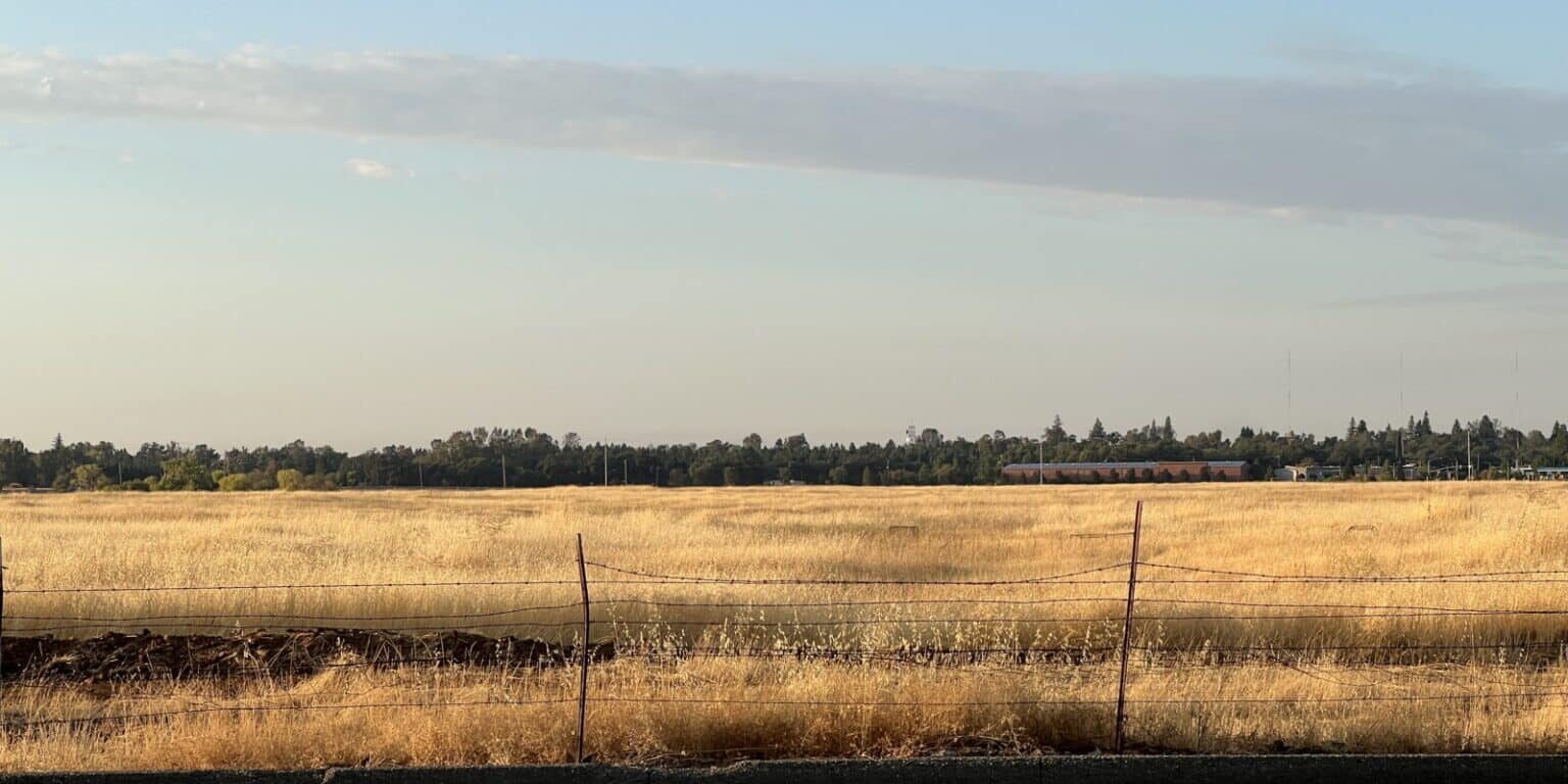 A barbed wire fence runs along the edge of a paved road, separating it from a dry grassy field under a clear sky—a tranquil scene perfect for travel inspiration or creative web design concepts, with trees and buildings visible in the distance.